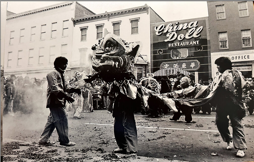 A black and white photo of a Lion Dance performance in front of China Doll Restaurant in Washington, D.C.'s Chinatown.