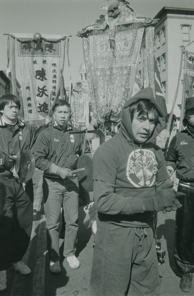 A man wearing a hoodie that reads "Jow Ga Kung Fu" stands with others holding signs as part of a parade.