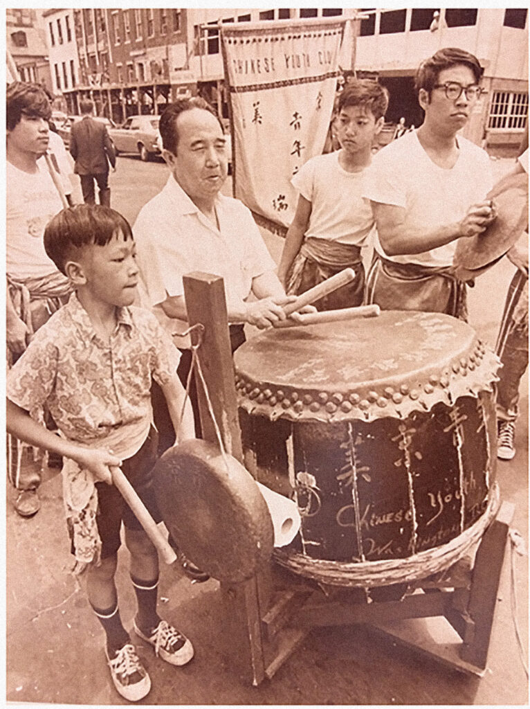 A group of people marching in a street parade with a large drum and gong.