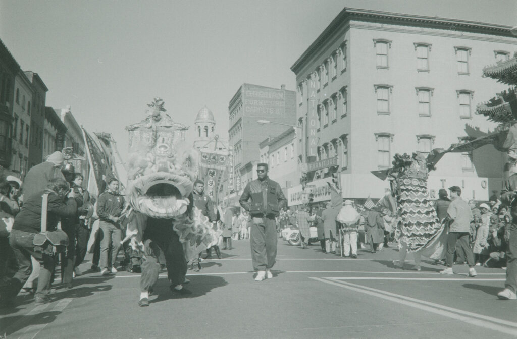 Two groups of Lion Dancers parade down a street while a man wearing sunglasses walks beside one of the lions.