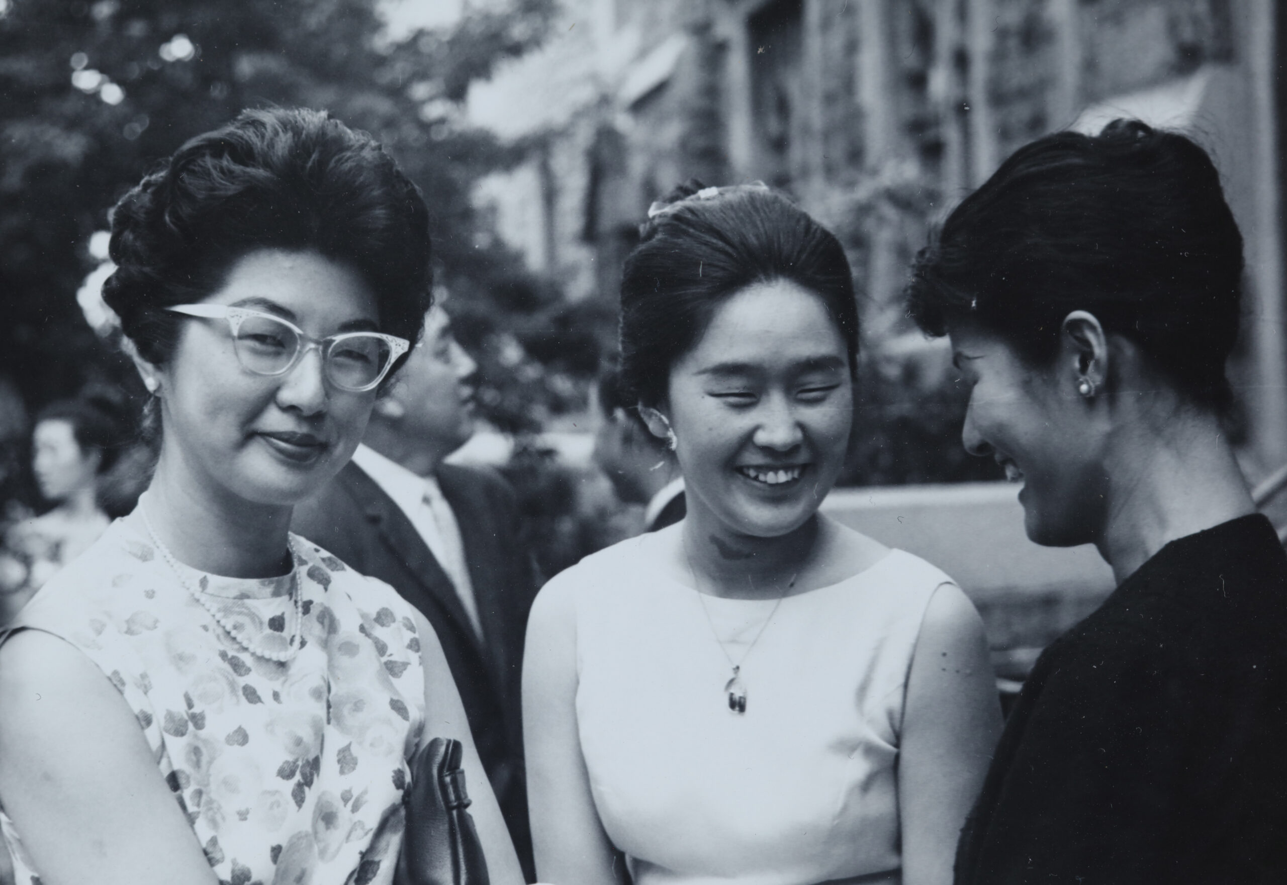 A woman in glasses and a sleeveless blouse smiles for the camera next to two friends smiling at each other.