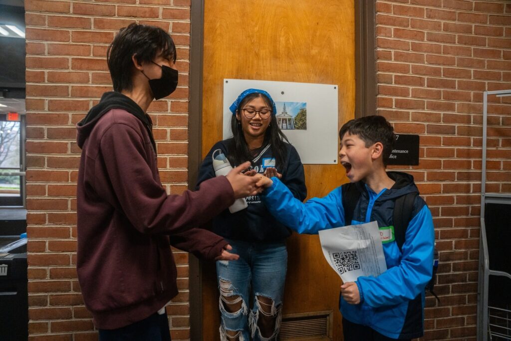 A youth wearing a maroon hoodie and a face mask greets a friend wearing a blue jacket while another friend smiles.