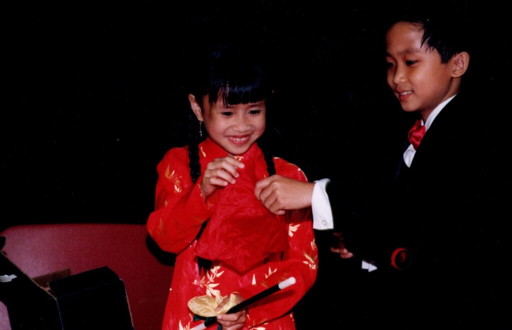 A young boy dressed in a magician's suit performs a trick with handkerchiefs for a young girl in a red dress.