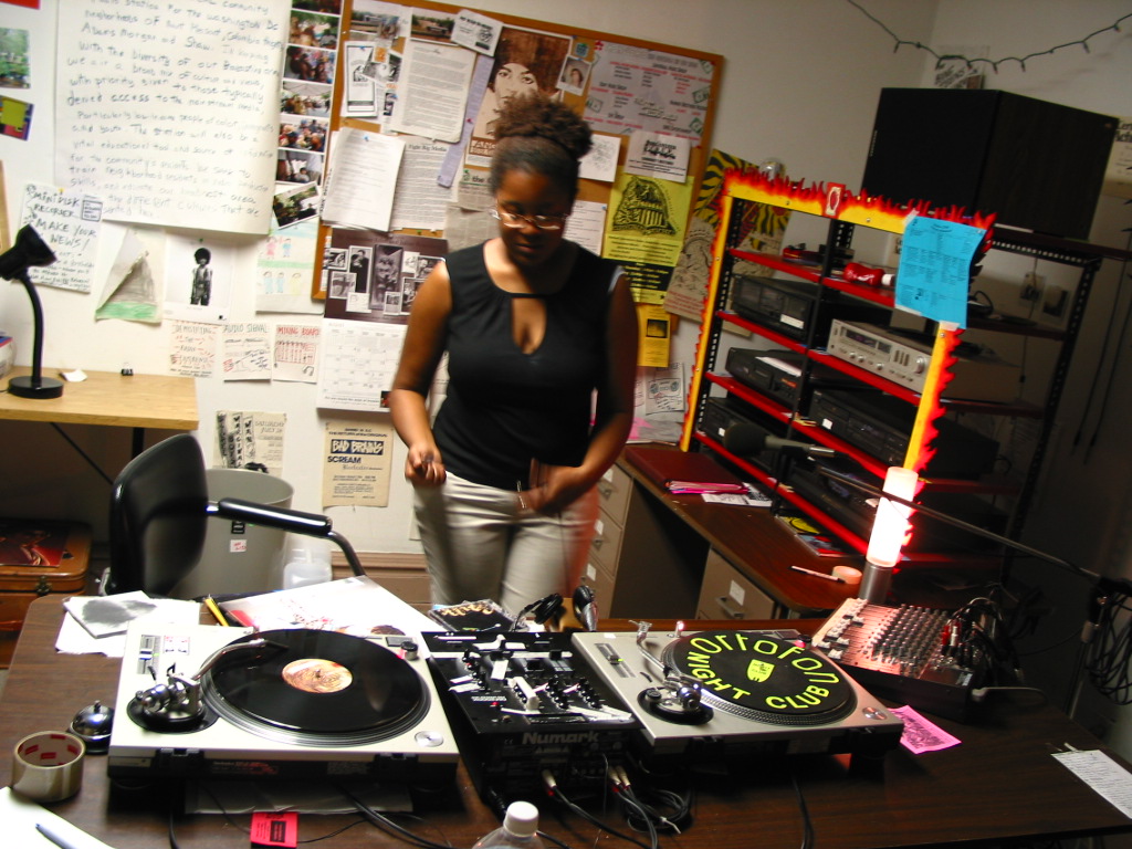 A DJ stands behind turntables. In the background is a bulletin board with flyers and an equipment rig.