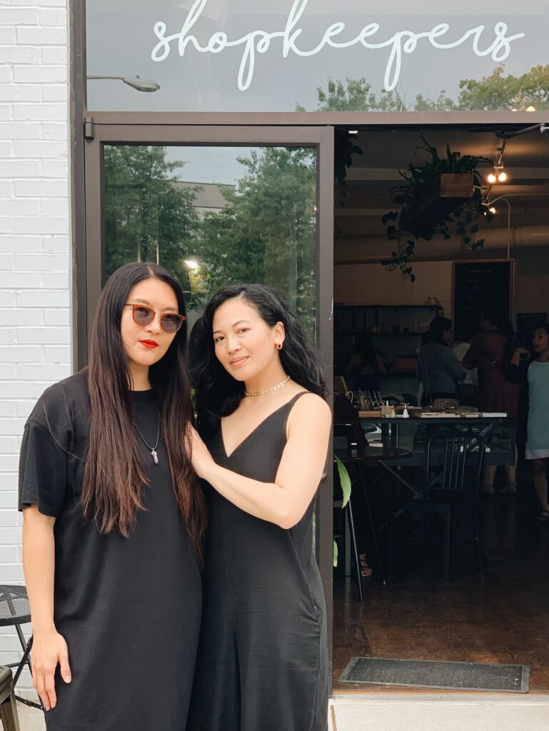 Two women wearing black outfits stand together in front of a building with a neon sign reading "shopkeepers."