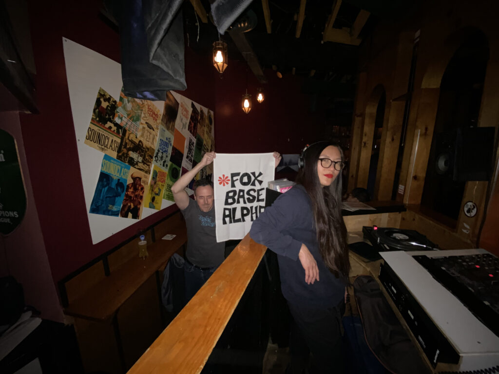 A DJ stands with her turntables while a man in the background holds up a flag that reads "Fox Based Alpha."