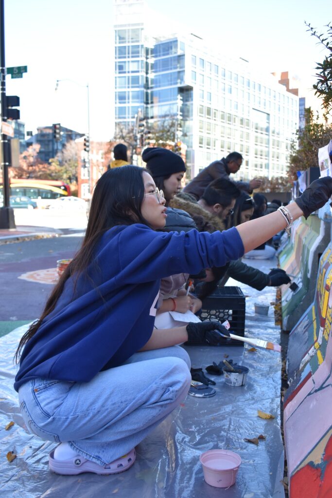 A young person wearing a blue sweatshirt and jeans helps paint a mural on a concrete traffic divider.