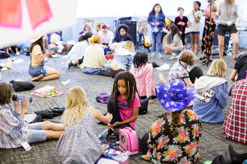 A large group of girls sitting throughout a classroom with gray carpet.