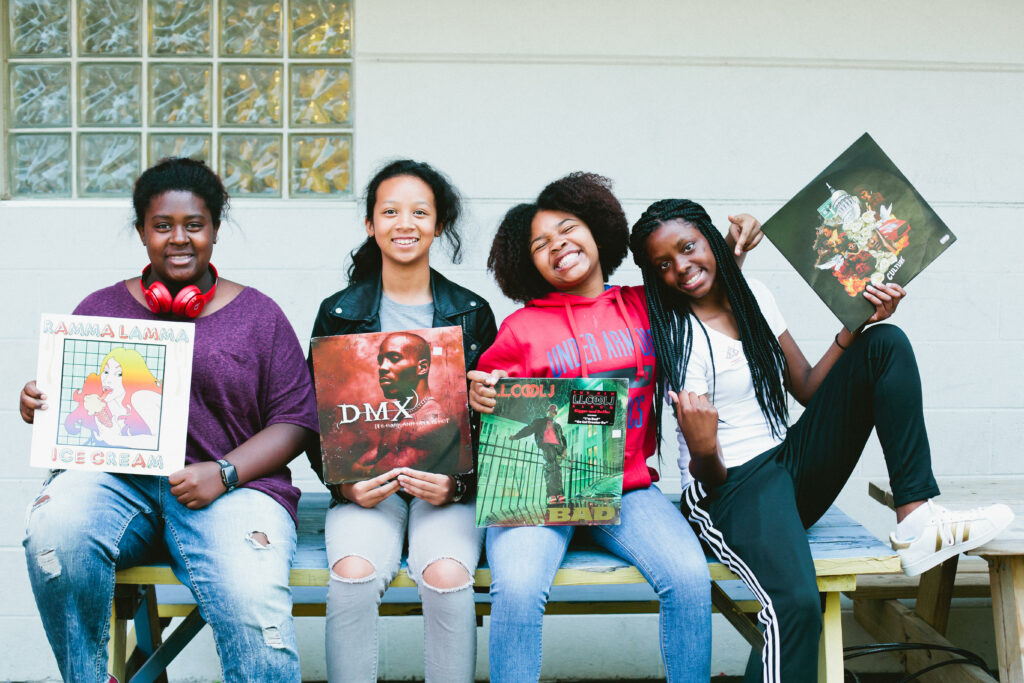 Four girls sit together to pose of a photo while smiling and holding vinyl record covers.