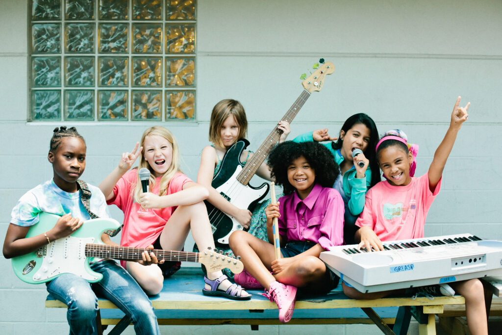 Six girls sit together on a bench and pose for a photo while holding guitars, microphones and a keyboard.