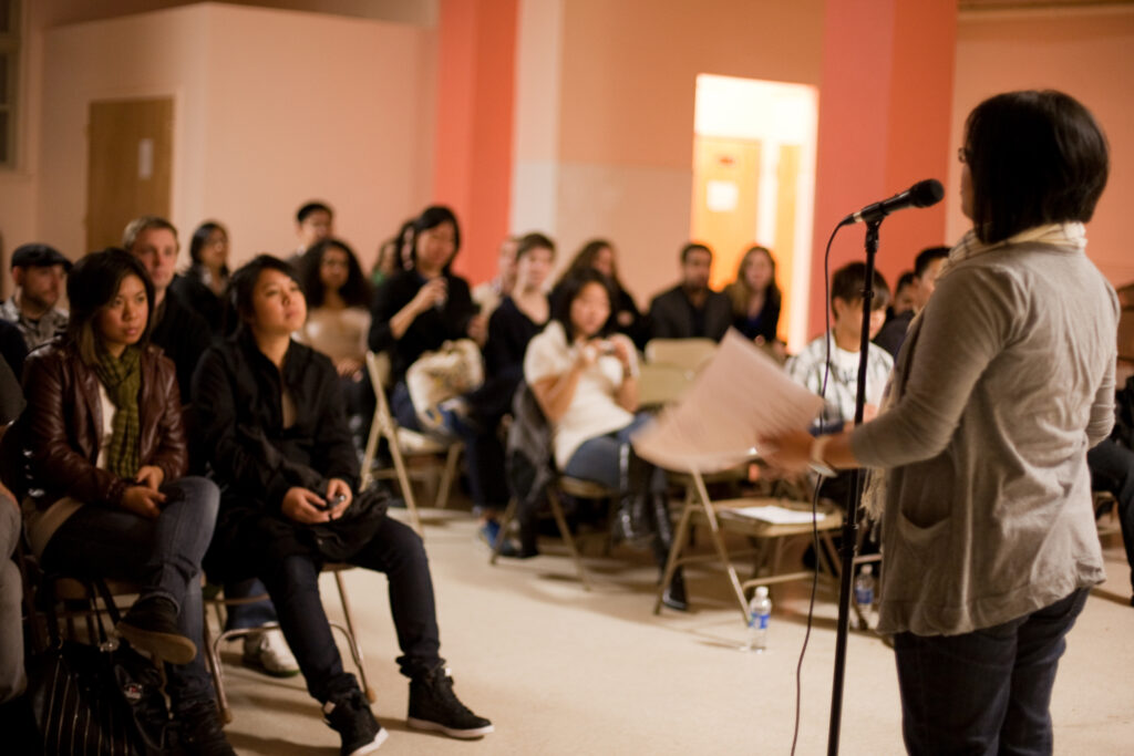 A performer stands in front of a microphone while an audience sits and listens.