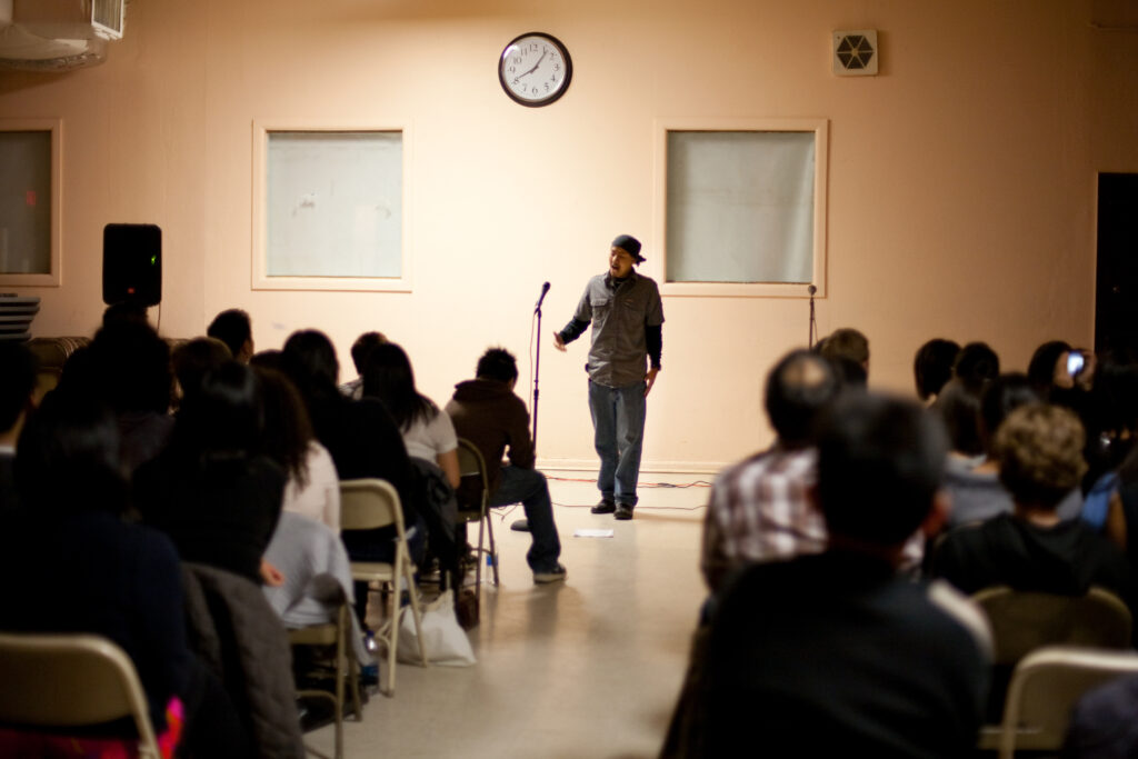 A shot from the back of the venue of a performer in front of a microphone speaking to a seated audience.