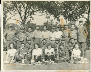 A black and white photo of a baseball team posing in a park in front of their trophies.